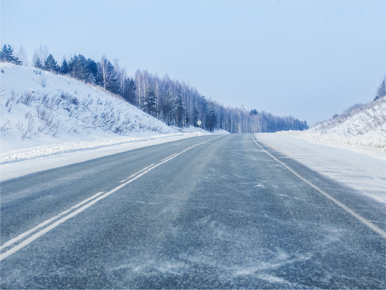 Snow drifts over a highway in winter framed on either side by snow covered hills and barren trees.