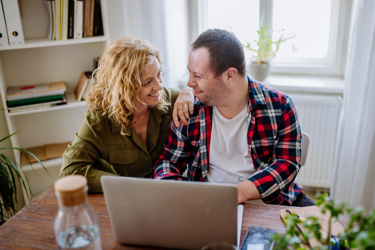 A young man with down syndrome works at his laptop with his mother.
